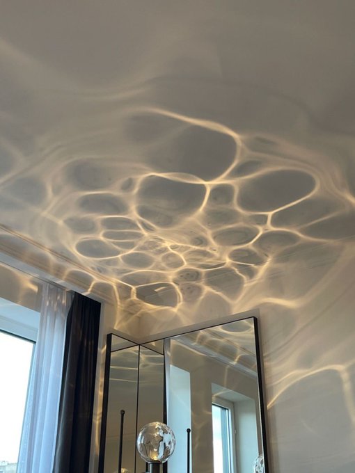 A photograph of a ceiling illuminated with a water caustics light effect, casting rippling patterns above a mirror and curtain in a modern room.