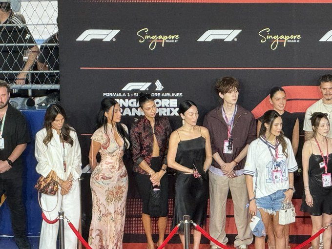 A group of people, including celebrities and event staff, posing on a red carpet at the F1 Singapore Grand Prix 2023, with the event branding visible on the backdrop behind them.
