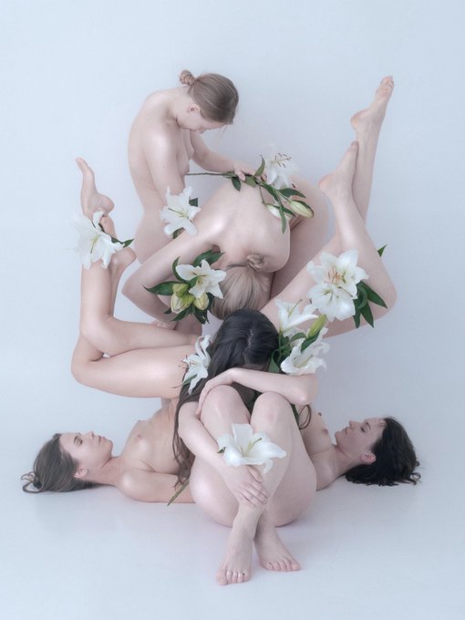 The image features a group of naked women lying down together, with white flowers surrounding them. There are four women in total, each one facing different directions. The flowers appear to be floating around the women, creating an ethereal atmosphere. The photo is titled "Bodies" and it seems to be part of a series or collection of images that explore themes related to human bodies and their connection with nature.