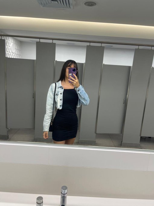 A woman takes a mirror selfie in a public restroom, wearing a black dress and a light blue denim jacket, holding a purple phone.