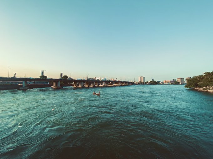 A scenic view from the water showing kayakers paddling on a turquoise sea, with a long bridge stretching across the horizon and city buildings visible in the distance under a clear blue sky.