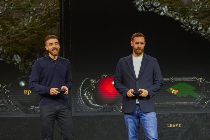 Two men stand on a stage holding game controllers in front of a large screen displaying a video game with a red orb; the text '2p' and a 'LEAVE' button are visible on the screen.