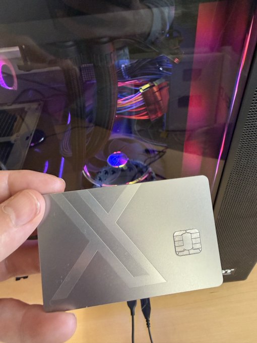 A hand holds up a silver metallic card with an embossed X design and a chip, held in front of a gaming PC case with colorful RGB lighting visible through the tempered glass side panel.
