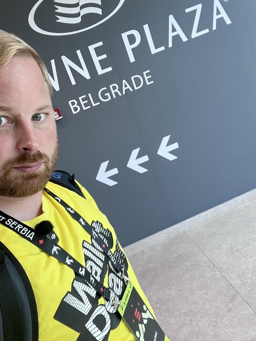 A man with a beard takes a selfie while wearing a yellow Waldom DAO shirt, standing near a sign reading 'WNE PLAZA BELGRADE.'