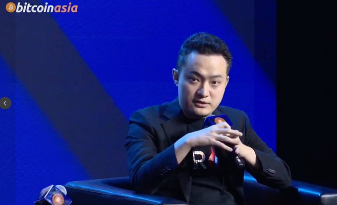 A man in a black suit speaks into a microphone labeled 'PN' on a stage with a blue background, with the 'bitcoinasia' logo visible in the top left corner.
