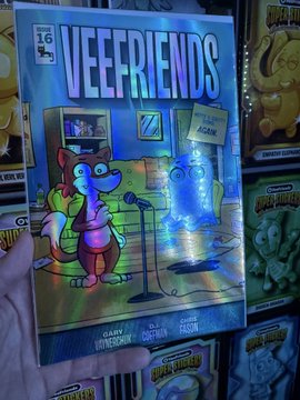 A photograph of a hand holding a holographic 'VEEFRIENDS' trading card (numbered 16) featuring an animal character on a stage, with other VeeFriends cards visible in the background.