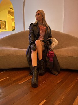 A woman with long hair wearing a black puffer jacket, striped top, and black boots, sitting on a beige couch in an indoor setting.