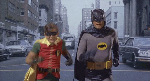 The image features two men dressed as Batman, one wearing a red cape and the other wearing a gray suit with a yellow bat symbol on his chest. They are walking down a street lined with cars parked along both sides of the road. The background includes buildings and trees, giving an urban setting to the scene. The caption "$useless and $fartcoin" is written in white text at the bottom of the image.