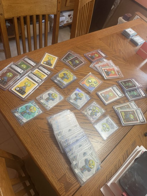 An overhead photo of a wooden dining table covered with numerous graded trading cards (mostly featuring Plants vs. Zombies characters) and several sealed card packs.