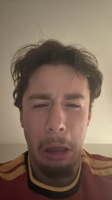 A close-up selfie of a man with messy brown hair and a striped red and yellow shirt making a pained or uncomfortable facial expression.