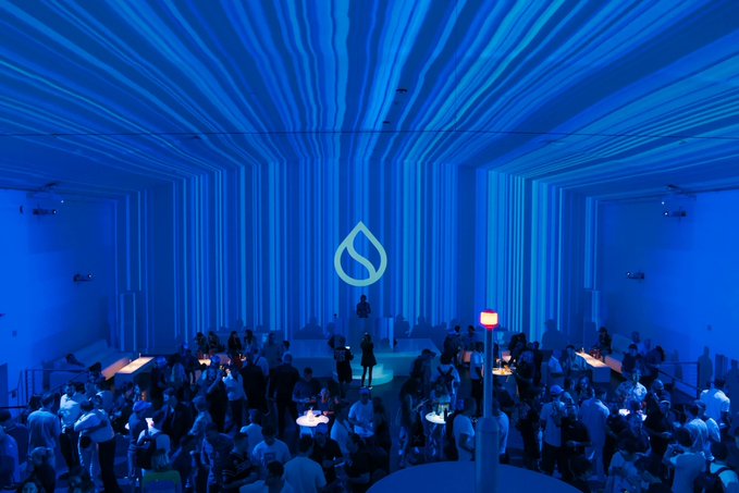 The image shows a large group of people gathered together, possibly at an event or party. The room has blue lighting that creates a unique atmosphere for the gathering. There are several tables set up with various items on them, including cups and bowls. A person is standing in front of one of these tables, likely overseeing the event or engaging with attendees. The image also contains text that reads "GM #Sui 生而不同", which could be a reference to the GM (General Motors) logo or a specific event related to GM's operations.