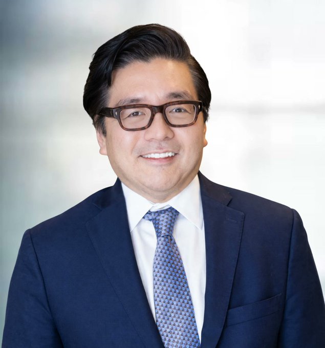 A professional headshot of an East Asian man wearing glasses, a navy blue suit, white shirt, and blue patterned tie, smiling at the camera against a blurred office background. The caption references a contrarian figure, with the image likely depicting a prominent crypto or financial figure—possibly Tom Lee of Fundstrat Global Advisors, given the @fundstrat mention in the post.