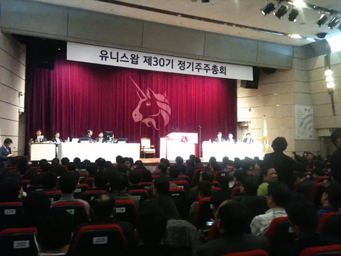 A large auditorium filled with attendees facing a stage with a red curtain and a unicorn logo; the banner reads '유니스왑 제30기 정기주주총회' (Uniswap 30th Regular General Meeting of Shareholders).