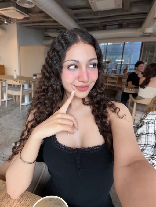 A selfie of a young woman with curly dark hair wearing a black top, posing with her finger near her face in a cafe or restaurant setting.