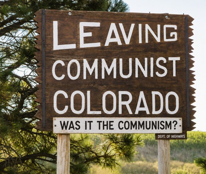 A wooden highway sign reading 'LEAVING COMMUNIST COLORADO - WAS IT THE COMMUNISM?' posted by the Dept. of Highways.