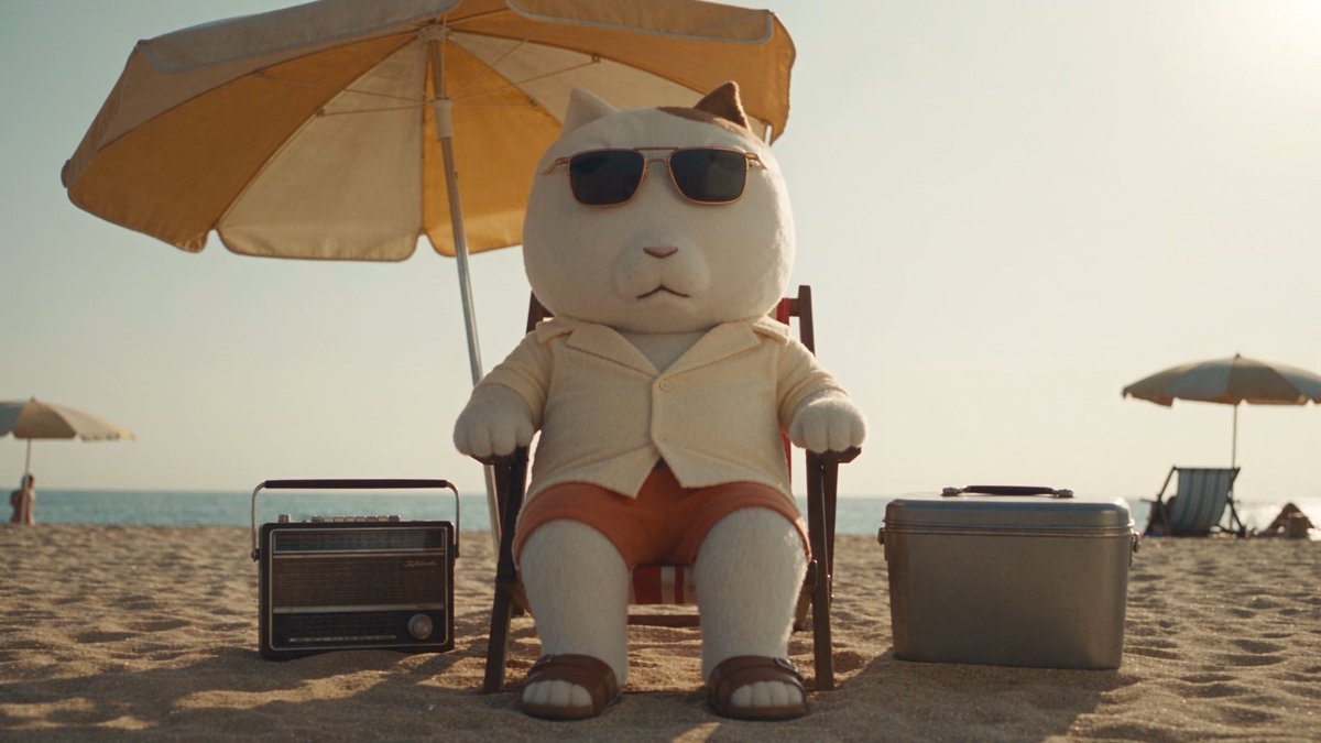 A cute white cat plush toy wearing sunglasses, a beige button-up shirt, and orange shorts sits on a beach chair under a yellow umbrella, flanked by a vintage radio and a cooler.
