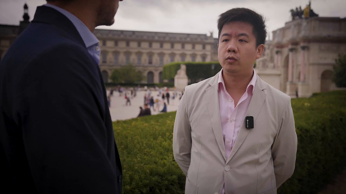 The image shows two men standing next to each other near a building with a large courtyard. One of them has his back turned towards the camera while the other man looks at him, possibly engaging in conversation or observing something interesting nearby. The scene takes place outdoors, and there are several people walking around in the background, indicating that it might be a popular location for visitors to explore.