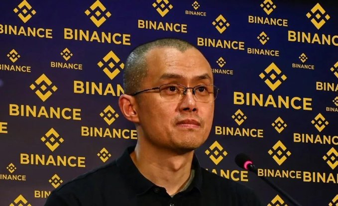 A close-up photo of Binance co-founder Changpeng Zhao (CZ) speaking in front of a dark backdrop covered in the yellow Binance logo.