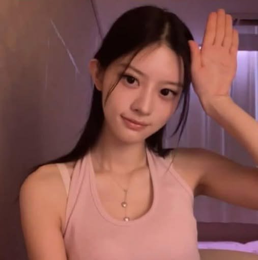 A young woman with long dark hair wearing a pink tank top poses with her hand raised near her face in a softly lit room.