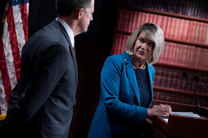 Two people at a formal event, with a woman in a blue blazer standing at a podium and a man in a grey suit beside her, framed by an American flag and shelves of books.