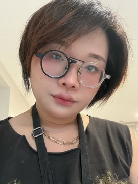 A woman with short brown hair and round black-rimmed glasses takes a selfie while wearing a black top and chain necklace, with "GM Artist /Art Qt yours" signaling a good morning greeting to her art community.