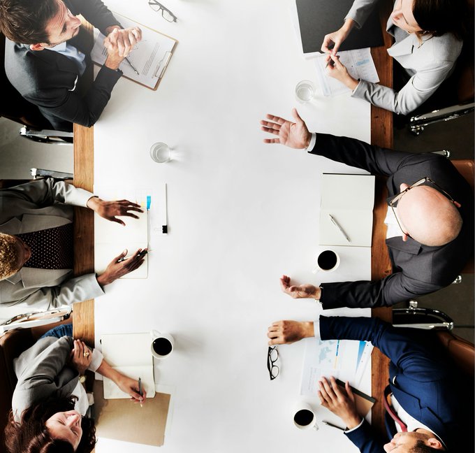 Six business professionals in suits are gathered around a white conference table viewed from above, gesturing and discussing with documents, notepads, and coffee cups in front of them. The image visually represents lawmakers and traders deliberating on legislation like the CLARITY Act, reflecting the political uncertainty behind Kalshi's under-25% odds on the digital assets bill passing by year-end.