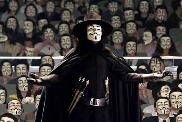 A figure wearing a black cloak, wide-brimmed hat, and a V for Vendetta mask stands with arms outstretched before a crowd all wearing identical masks.