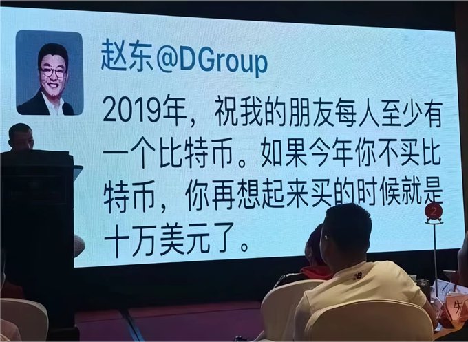 A photo of a presentation screen showing a quote from "赵东@DGroup" stating "2019年，祝我的朋友每人至少有一个比特币。如果今年你不买比特币，你再想起来买的时候就是十万美元了。" (In 2019, I wish my friends each have at least one Bitcoin. If you don't buy Bitcoin this year, when you think of buying it, it will be $100,000).