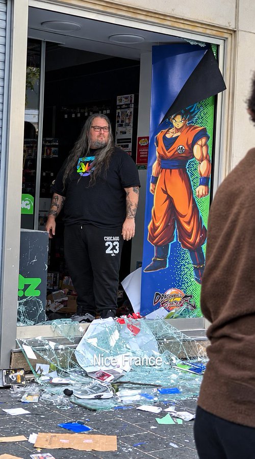 A man stands inside a damaged storefront amid shattered glass and debris, with a large Dragon Ball-style character poster beside him and the text “Nice, France” near the bottom.