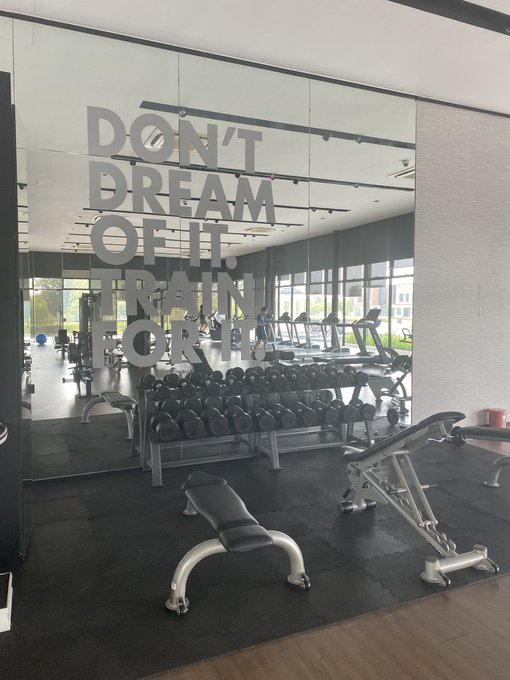 A gym interior featuring a large mirror with the motivational quote 'DON'T DREAM OF IT. TRAIN FOR IT.' applied to the glass, with dumbbells and workout benches visible.