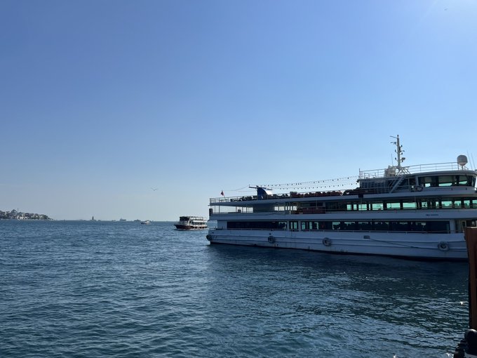 A large white passenger ferry boat on blue water under a clear sky, with a smaller boat in the distance and a coastline visible on the left.