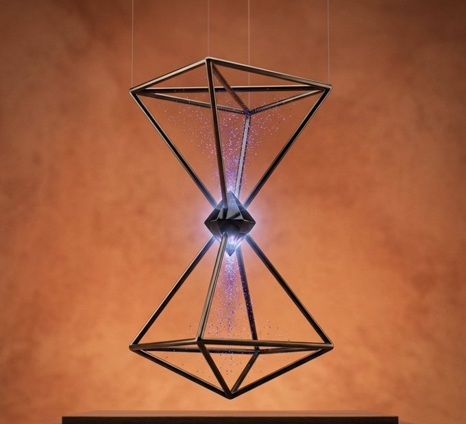 A geometric hourglass-shaped sculpture with dark metal frames and glowing pink/purple particles inside, displayed against a warm orange backdrop.