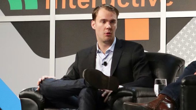 Man in dark suit and light blue shirt seated on stage at what appears to be a TechCrunch Disrupt interview, with the event's colorful geometric backdrop visible behind him.