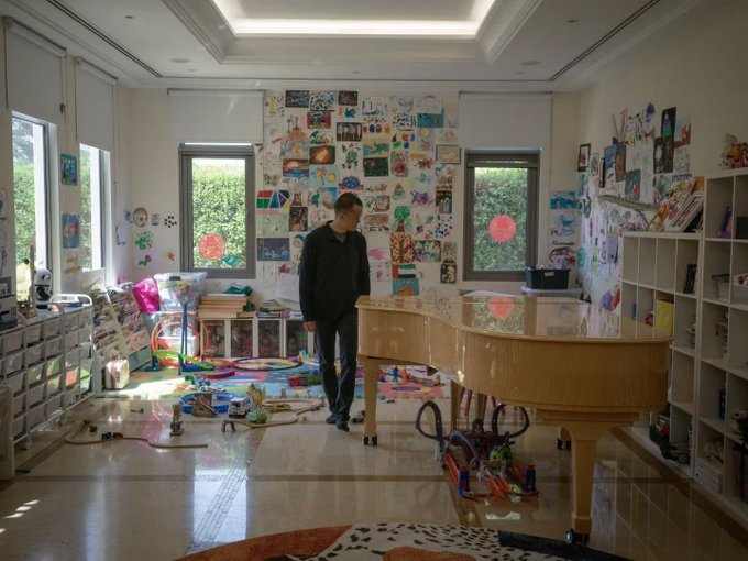 A man stands in a bright, art-filled room next to a grand piano, surrounded by children's drawings, colorful toys, and creative supplies scattered across the floor.

The image shows the interior of a spacious, well-lit home filled with children's artwork covering the walls, a grand piano, and various toys scattered across the polished floor, while a man in dark clothing stands looking downward. The post about CZ Binance buying a house seems unrelated to this domestic scene, which appears to depict a family home or art studio rather than anything cryptocurrency-related.