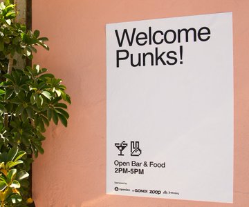 The image features a white sign with black text that reads "Welcome Punks!". The sign is attached to a pink wall, which has some greenery growing on it. There are also two potted plants visible in the background - one near the left side of the image and another towards the right side. The main focus of the image is the white sign with black text that reads "Welcome Punks!".