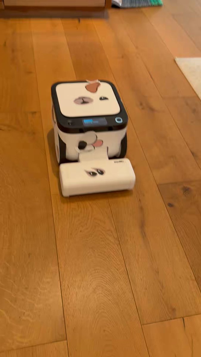 The image shows a white robot vacuum cleaner with black and red accents sitting on a wooden floor. The robot has a face, which appears to be a cat or dog, giving it a playful appearance. In the background, there are two books placed next to each other, possibly containing information about the robot's features or instructions for use.