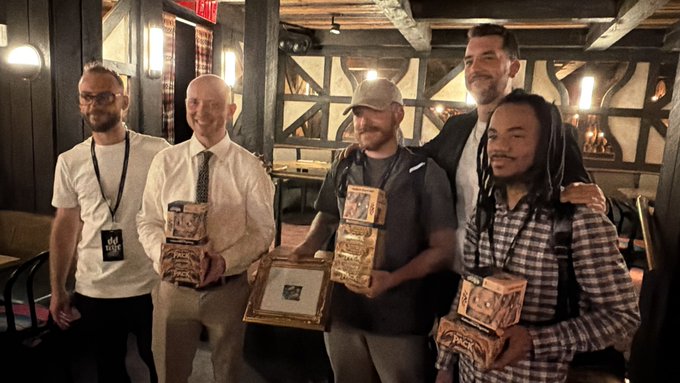 A group photo of five men standing together in what appears to be a rustic tavern or pub, with three of them holding ornate wooden/decorative boxes or plaques.