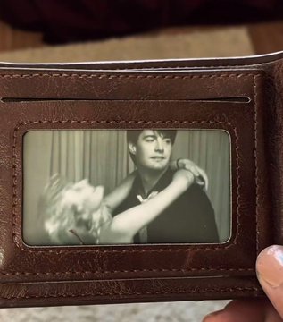 A hand holds open a brown leather wallet to reveal a black-and-white photo ID of a young man holding a small dog.