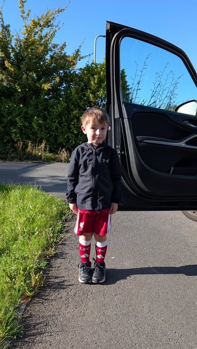 A young boy in a black Under Armour jacket, red shorts, and tall socks stands on a paved road next to an open car door.