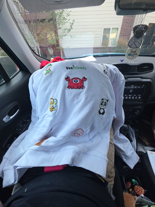 A white 'Veefriends' hoodie with various embroidered cartoon characters lies on the passenger seat of a car, with a pug air freshener hanging from the rearview mirror.