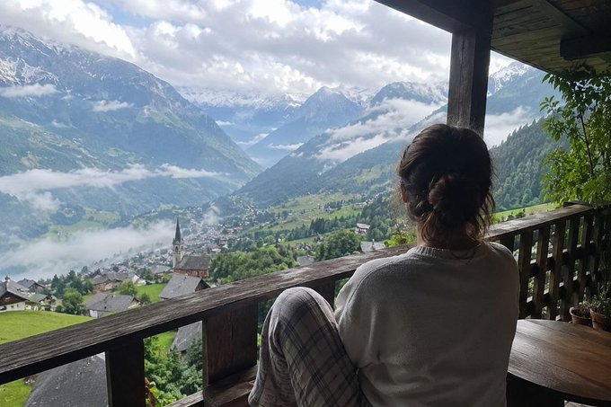 A woman sits on a wooden balcony overlooking a picturesque Alpine valley with a village, church steeple, green meadows, and snow-capped mountains partially covered in clouds.