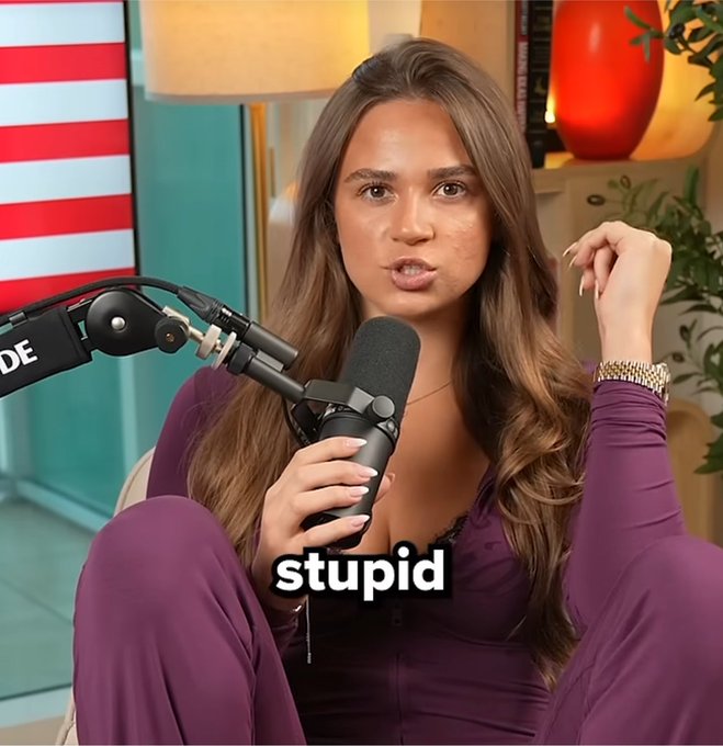 A woman with long brown hair, wearing a purple top, holds a microphone with "DE" visible on it while gesturing with her raised hand, with the word "stupid" displayed as a caption. The post's question "Who sits like this on air" appears to reference her unusual seated posture, framing the clip as a humorous critique often used in crypto/memecoin promotional content.