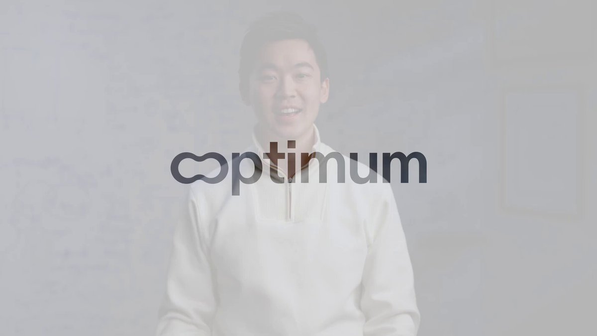 The image shows a man wearing a white shirt with the word "Optimum" written across his chest. The man appears to be smiling, possibly posing for a photo or video. The background of the image features a brick wall, which could be part of an office space or a room in a building.