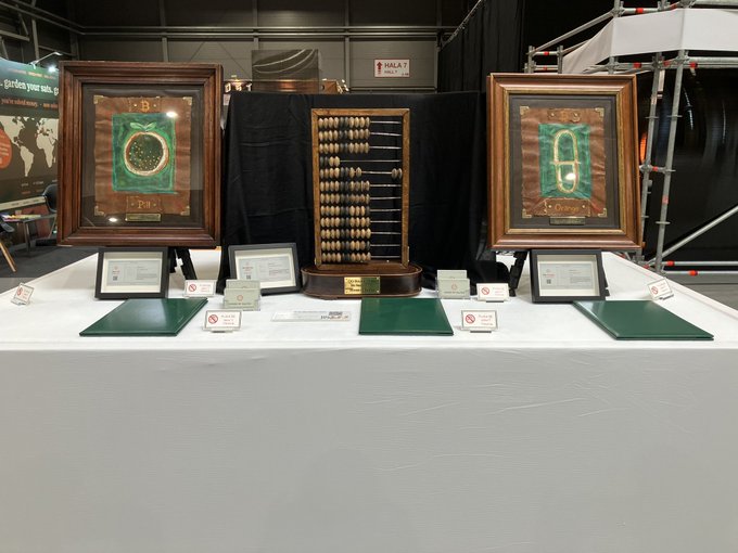 The image shows a table at an art gallery displaying three framed pictures of clocks. The first clock has a green frame, the second one has a brown frame, and the third one also has a green frame. Each picture is accompanied by a plaque that reads "There is no words to describe, you need to see it with your eyes Come to art gallery to witness it @BTCPrague". This arrangement suggests that these clocks are part of an exhibition or showcase at the gallery, where visitors can appreciate and learn about the different styles and designs of the clocks.