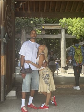 A young man in a white t-shirt and jean shorts stands with his arm around a blonde woman wearing a yellow dress, appearing to pose closely together in what looks like a temple or shrine courtyard. The image plays on the phrase "airdrop," humorously suggesting an "airless drop" since the man appears to be withholding affection, keeping his arm positioned away from a full embrace.