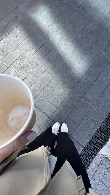 A first-person perspective photo showing a person holding a paper cup of coffee, looking down at their white sneakers and black pants on a paved outdoor surface.