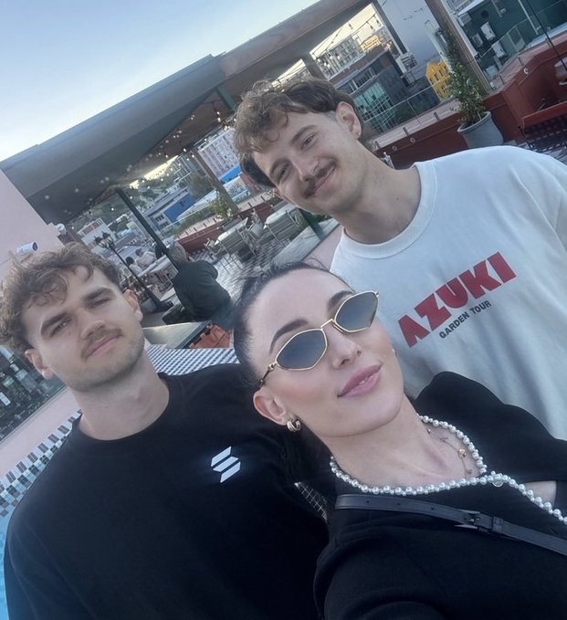 A selfie of three people on an outdoor patio; one man wears a black shirt and another wears a white shirt with 'AZUKI GARDEN TOUR' text, while a woman with sunglasses and a pearl necklace is in the foreground.