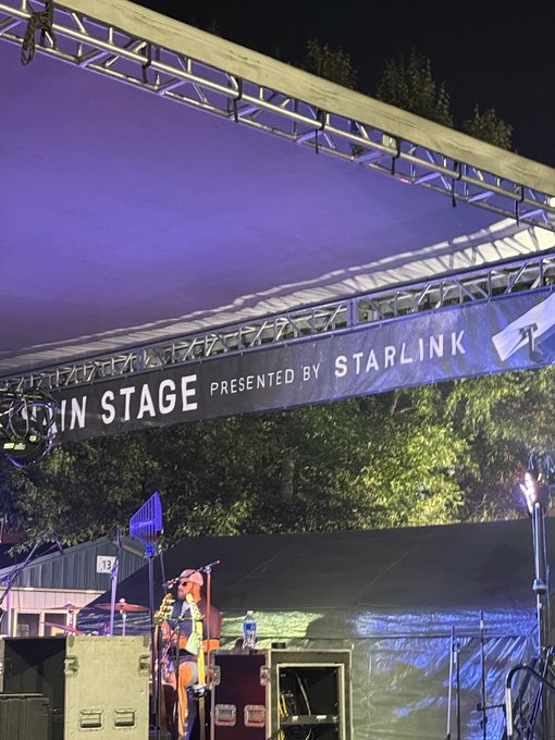 A nighttime view of an outdoor concert stage with a banner reading 'MAIN STAGE PRESENTED BY STARLINK', showing a performer with a guitar under purple lighting.