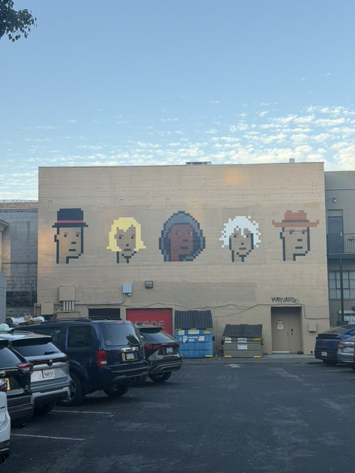 A pixel-art mural of five interchangeable faces with different hats adorns a building wall, with "MURVAIR" tagged on the door. The comment "Extremely fungible" plays on the mural's depiction of nearly identical faces that can be swapped out like interchangeable tokens.