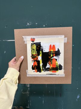 A hand holding up a small abstract painting mounted on a square piece of brown cardboard, featuring black, white, red, yellow, and green shapes against a dark green wall.